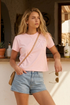 Woman wearing a pink t-shirt with bow fabric design and denim shorts standing in a sunlit in front of a cantina. 