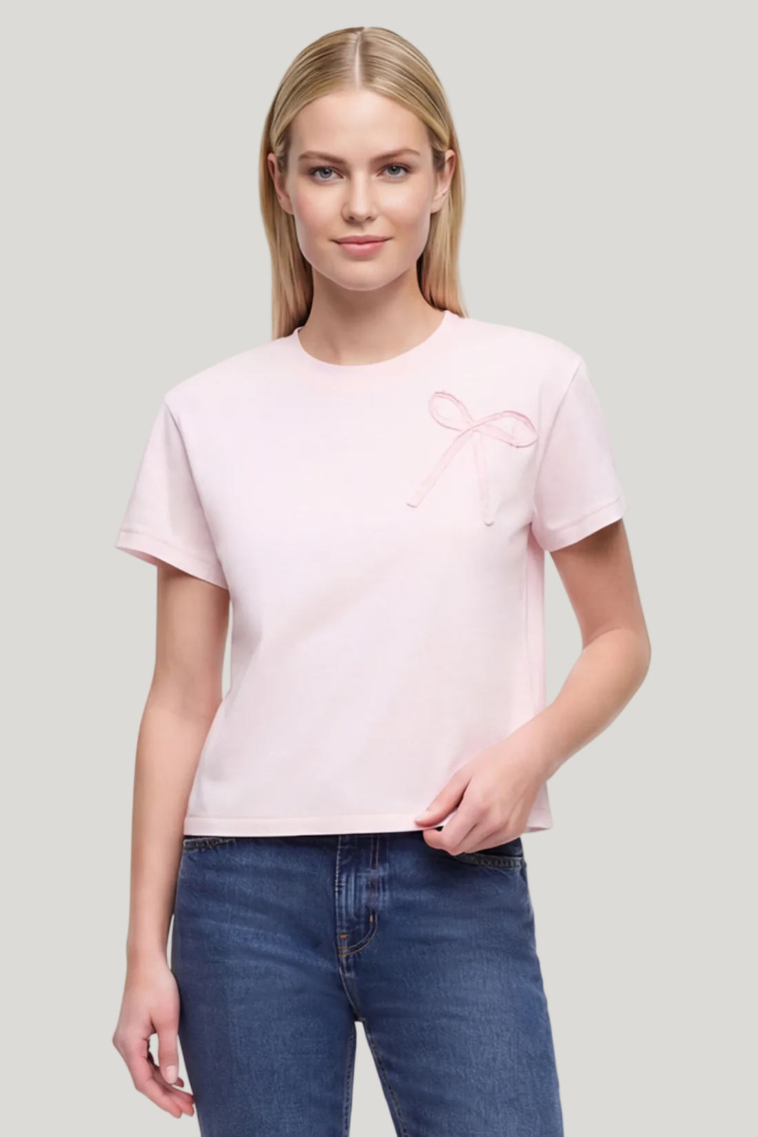 Front view of woman wearing a light pink t-shirt with a bow fabric design and blue jeans on a white background. 