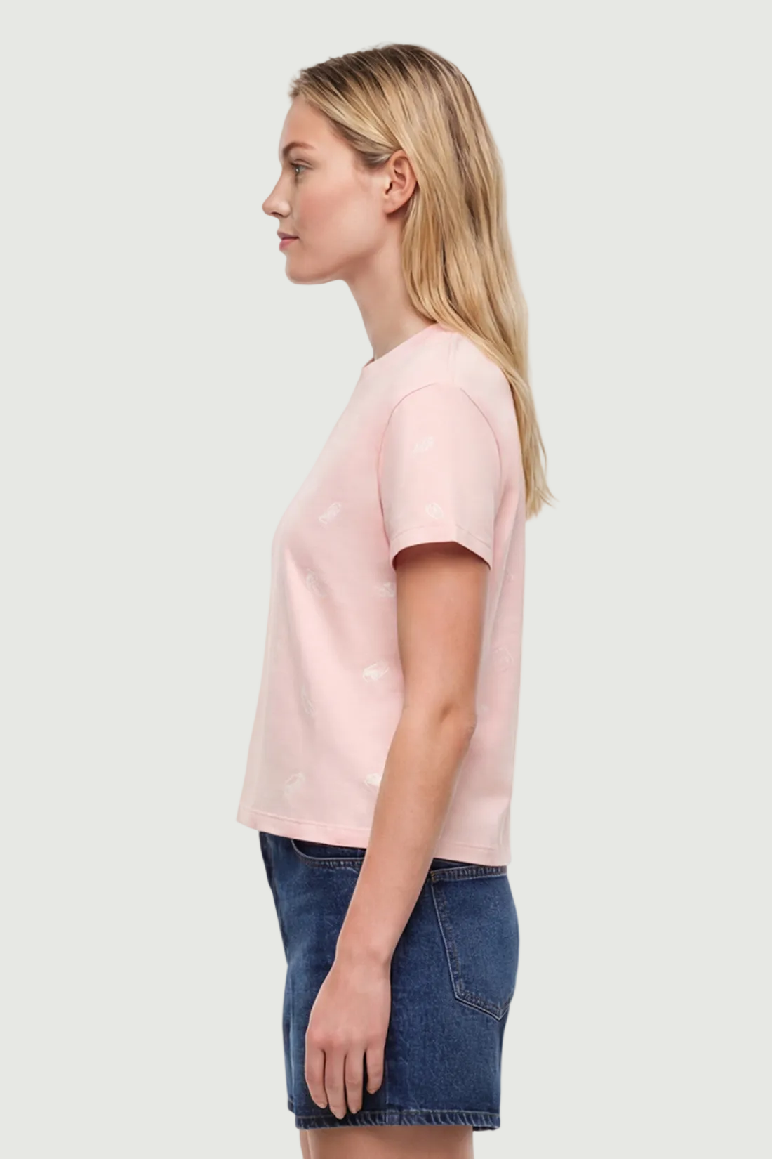 Side view of woman wearing a light pink t-shirt with pasta noodle shapes  pattern design and denim shorts on a white background. 