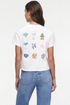 A woman viewed from the back wearing a white short-sleeve crop top T-shirt with a Mahjong grid of colorful cocktail-themed illustrations on the back, paired with blue jeans, against a plain white studio background. 