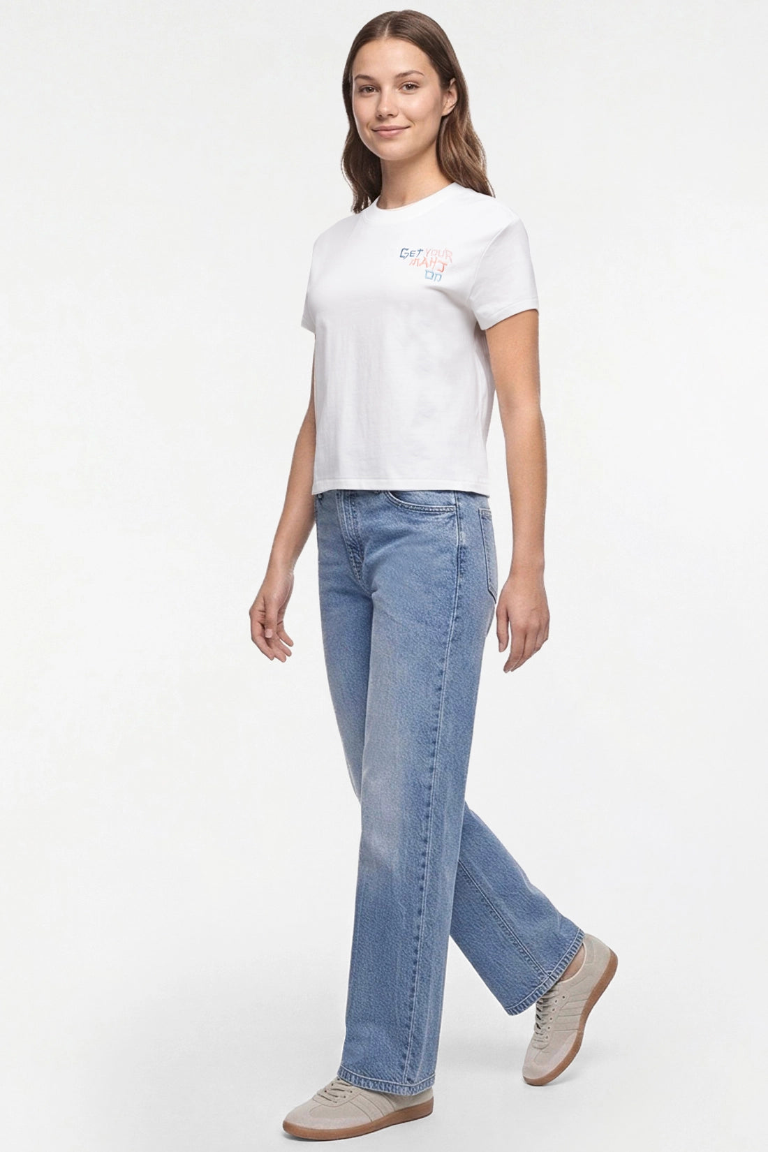 A woman modeling a white short-sleeve crop top with a small pastel graphic on the chest, paired with light-wash blue jeans and beige sneakers, against a plain white studio background. 