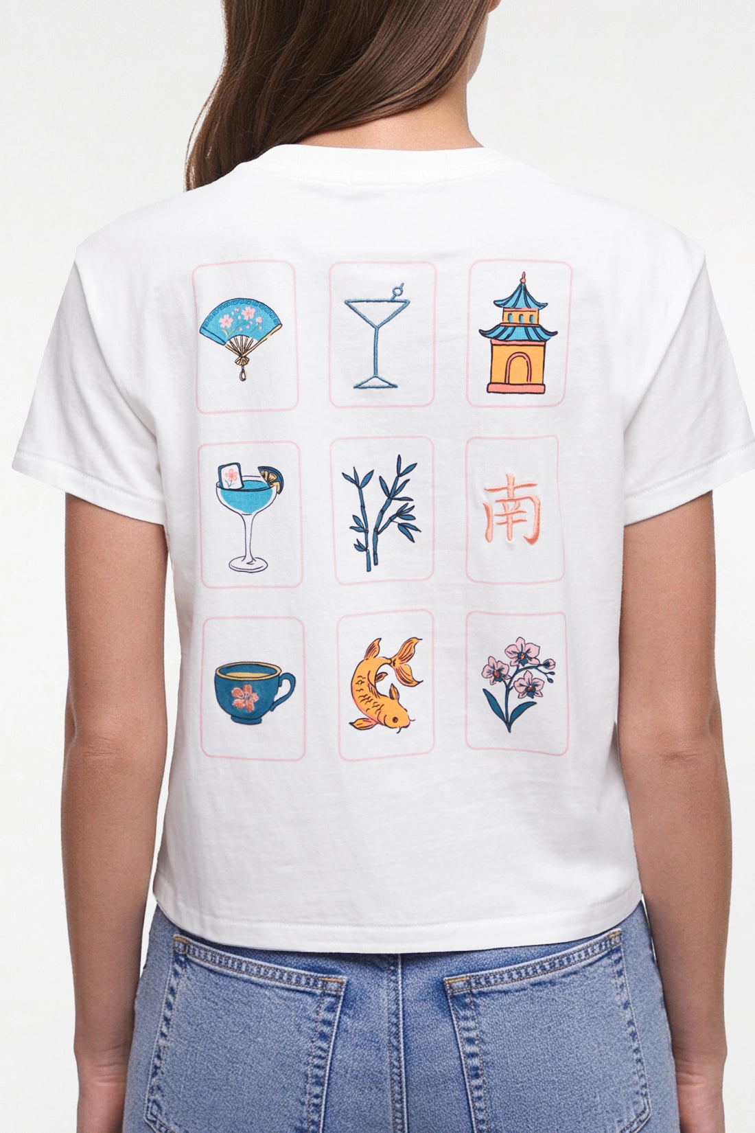 A woman shown from the back wearing a white short-sleeve T-shirt with a Mahjong grid of nine colorful illustrated icons on the back, paired with blue jeans against a plain white studio background.  