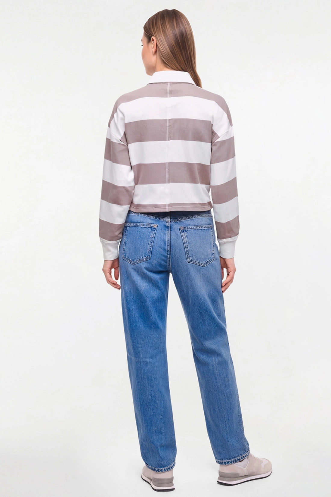 A woman shown from the back wearing a cropped long-sleeve collared shirt with horizontal taupe and white stripes, paired with blue jeans and light gray sneakers, against a plain white studio background. 