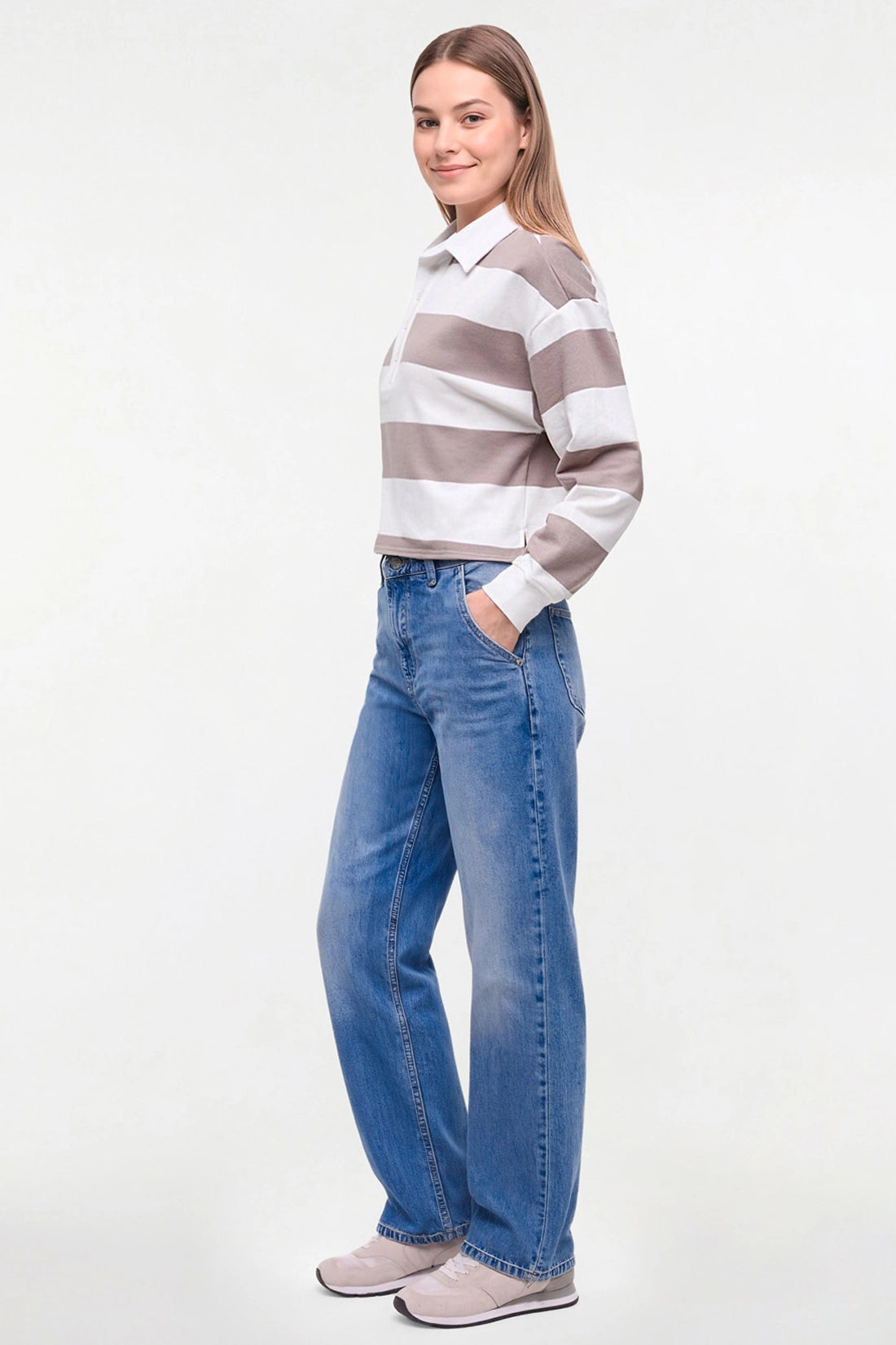 A woman modeling a long-sleeve collared knit top with horizontal taupe and white stripes, paired with blue jeans and light gray sneakers, against a plain white studio background. 