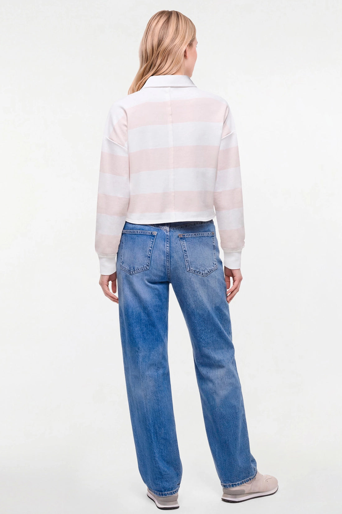 A woman viewed from behind wearing a pale pink and white horizontally striped long-sleeve crop top with blue jeans and gray slip-on shoes, against a plain white studio background. 