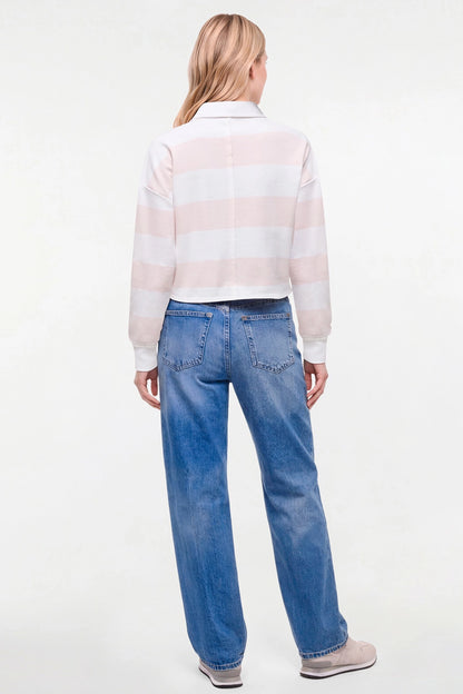 A woman viewed from behind wearing a pale pink and white horizontally striped long-sleeve crop top with blue jeans and gray slip-on shoes, against a plain white studio background. 