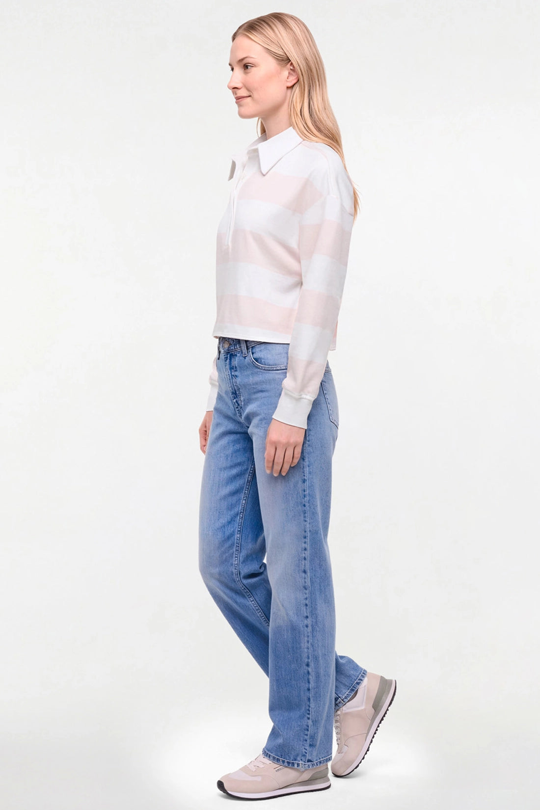 A woman in profile wearing a light pink and white striped knit long-sleeve shirt, light-wash blue jeans, and pale pink sneakers, standing against a plain white studio background. 