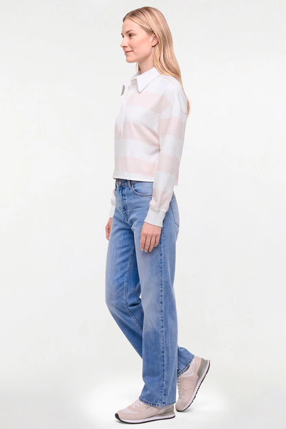 A woman in profile wearing a light pink and white striped knit long-sleeve shirt, light-wash blue jeans, and pale pink sneakers, standing against a plain white studio background. 