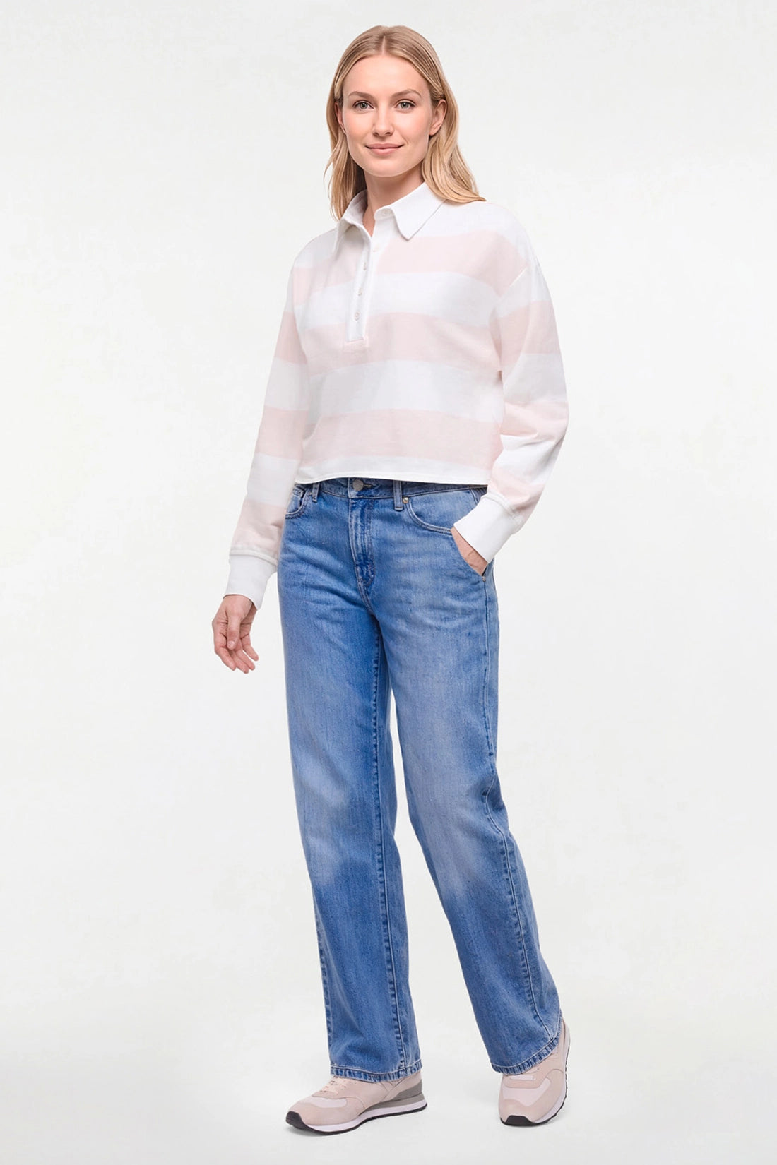 A woman modeling a white and pale pink striped long-sleeve polo shirt with blue jeans and light-colored sneakers, standing against a plain white studio background. 