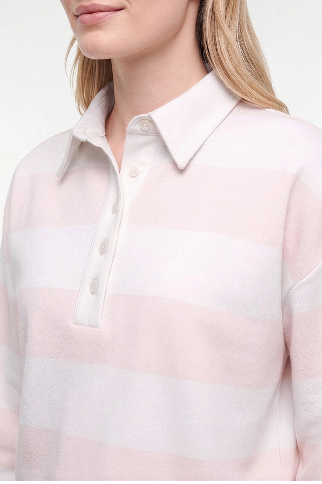 A close-up of a model wearing a pale pink and white horizontally striped knit polo shirt with a collar and three-button placket. 