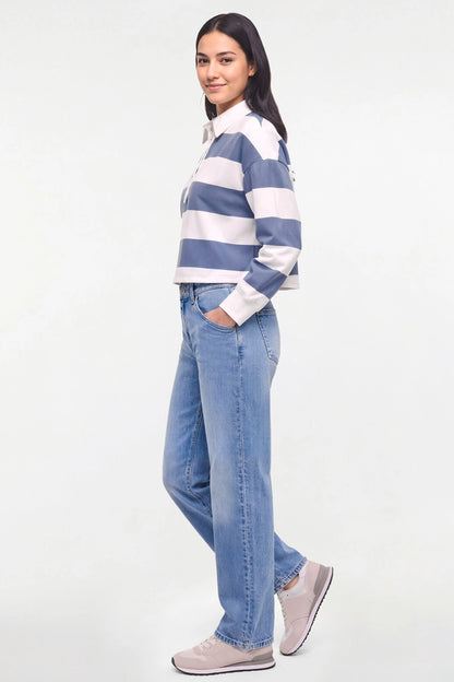 A woman modeling a blue and white striped long-sleeve rugby shirt with light-wash blue jeans and light gray sneakers, standing against a plain white studio background. 