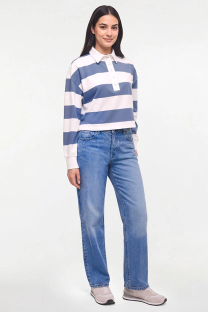 A woman modeling a blue and white striped long-sleeve rugby shirt with light-wash blue jeans and gray sneakers, standing against a plain white studio background. 