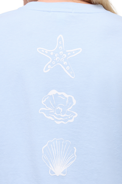 Close up view of light blue t-shirt with white starfish, shell, and pearl designs on a plain background. 