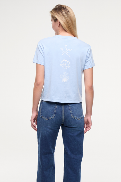 Back view of woman wearing light blue t-shirt with white starfish, shell, and pearl designs and blue jeans on a plain background. 