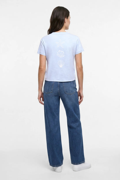 Back view of woman wearing light blue t-shirt with white starfish, shell, and pearl designs and blue jeans on a plain background. 