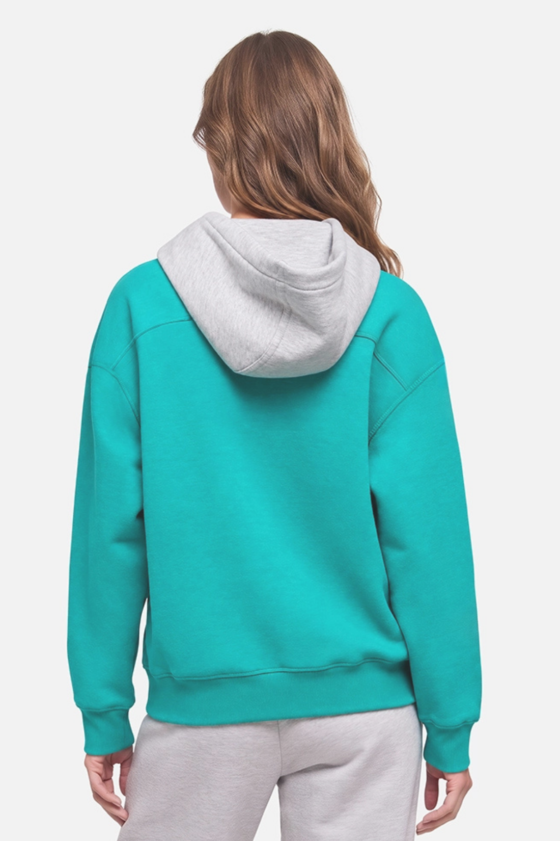 A woman viewed from the back wearing a teal-green and light gray hooded sweatshirt, paired with light gray sweatpants, against a plain light gray studio background. 