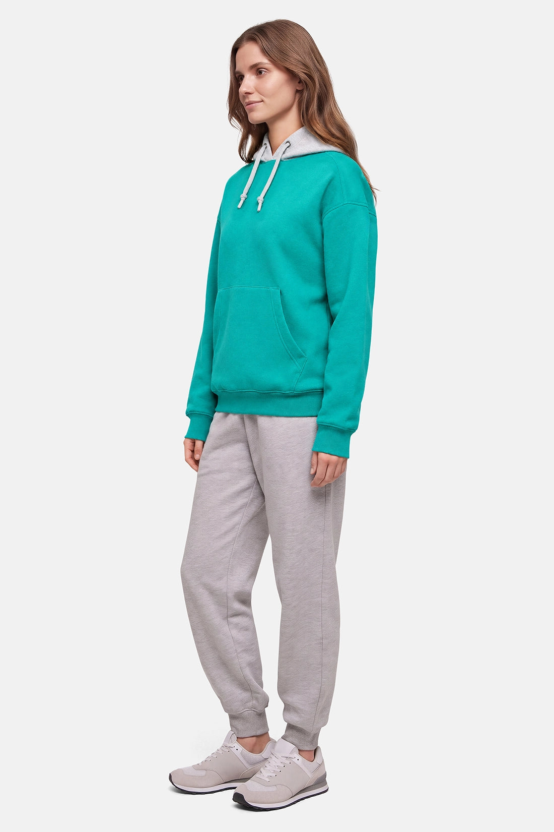 A woman modeling a teal green pullover hoodie with white drawstrings and light gray jogger sweatpants, paired with gray sneakers, against a plain light gray background. 