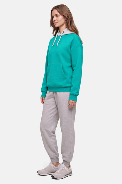 A woman modeling a teal green pullover hoodie with white drawstrings and light gray jogger sweatpants, paired with gray sneakers, against a plain light gray background. 