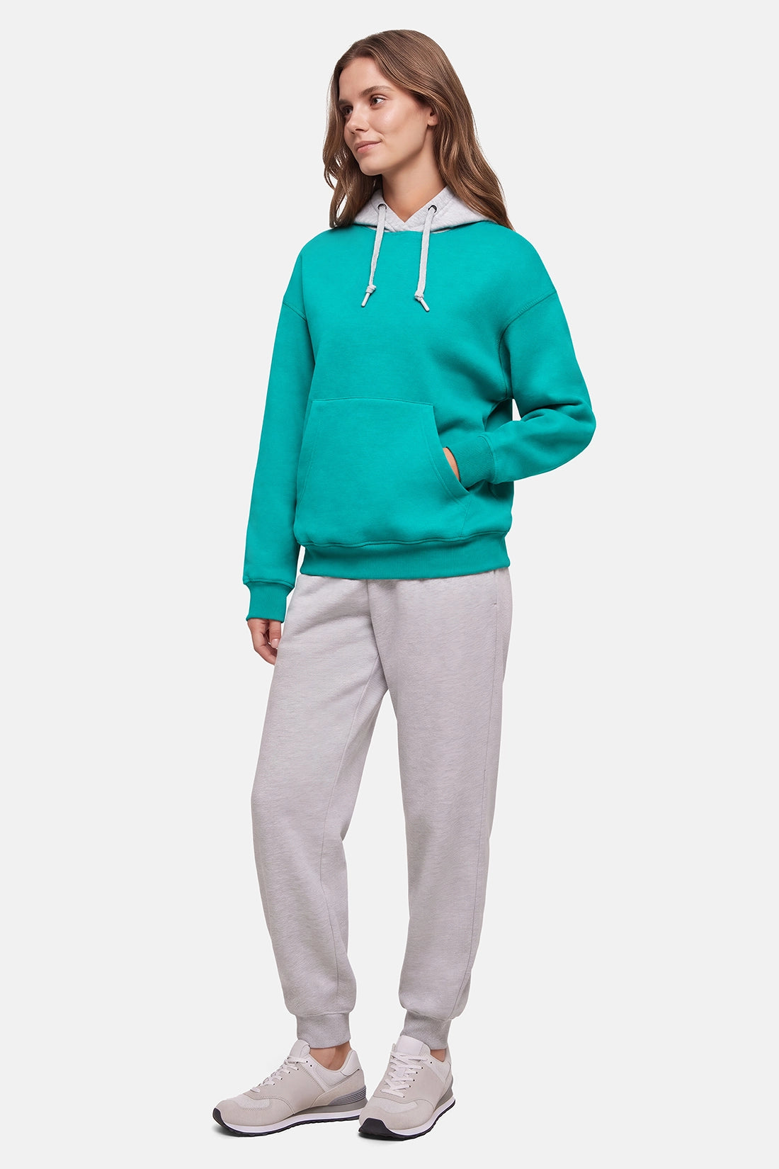 A woman modeling a bright teal-green hoodie with a white inner collar and white drawstrings, paired with light gray jogger pants and white sneakers, against a plain light gray studio background. 
