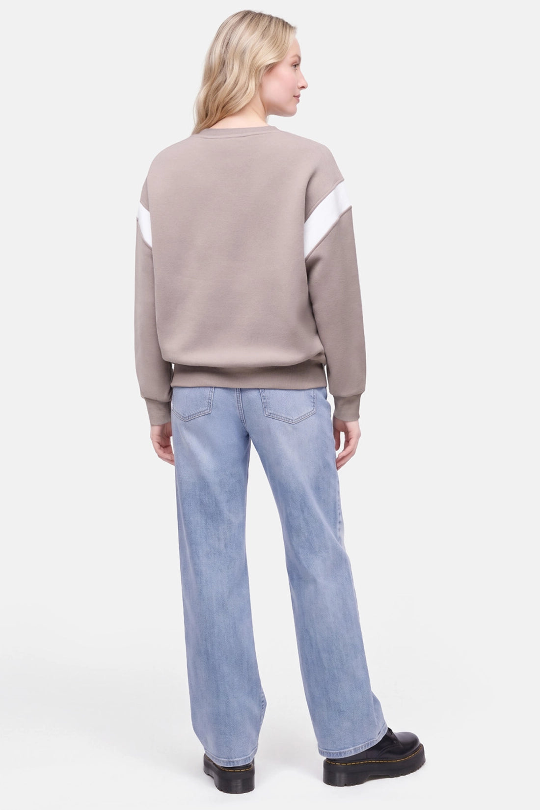 A woman shown from the back wearing a taupe crewneck sweatshirt with white diagonal sleeve stripes, light blue jeans, and black loafers, against a light gray studio background. 
