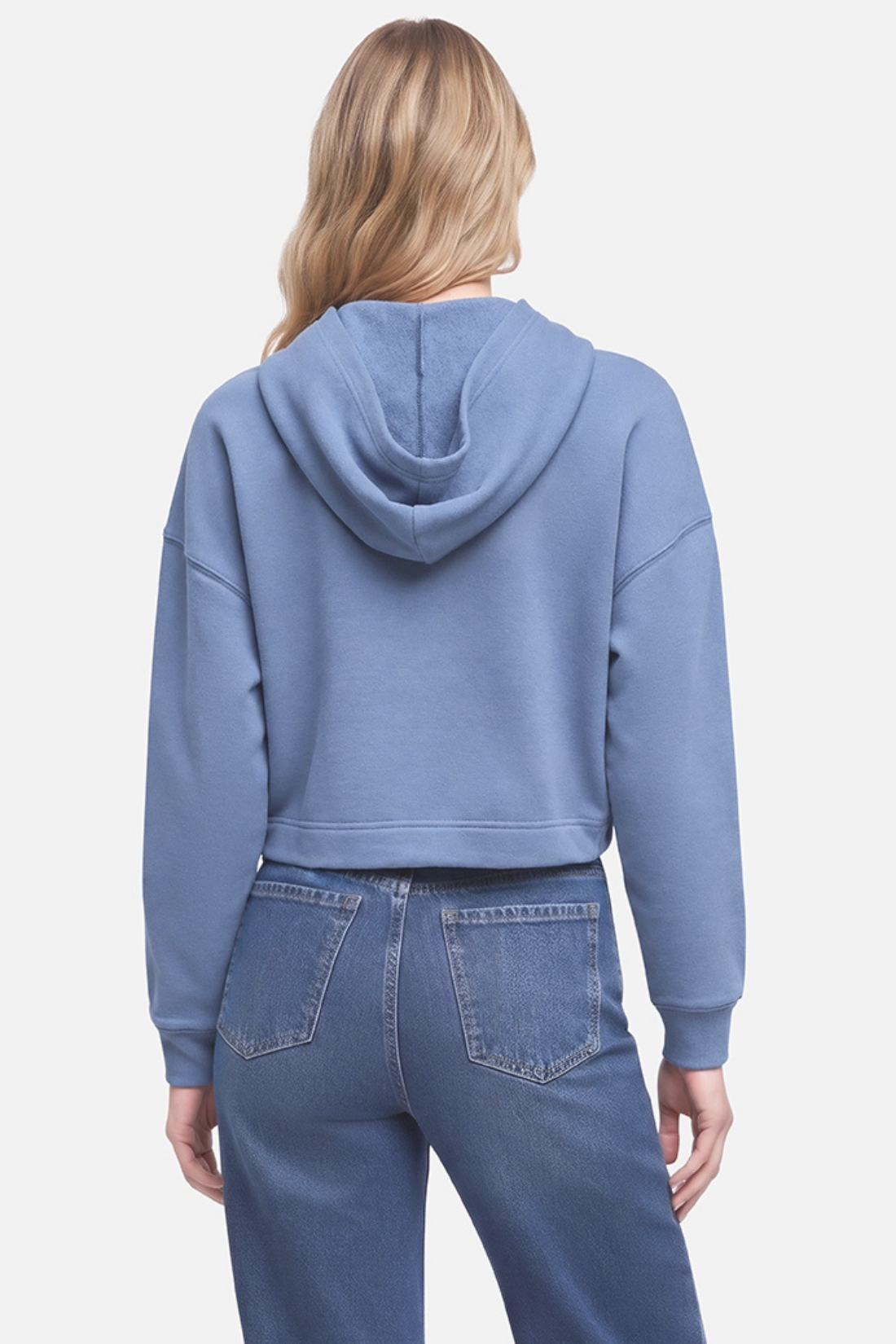 A woman shown from the back wearing a dusty blue cropped hoodie and blue jeans against a light gray studio background.