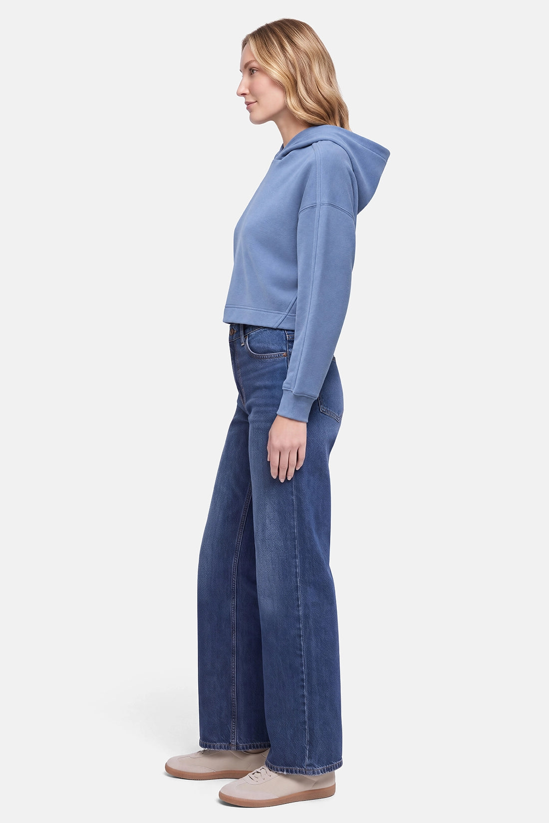 A woman shown in profile wearing a light blue cropped hoodie, dark blue wide-leg jeans, and beige shoes against a plain light gray studio background.
