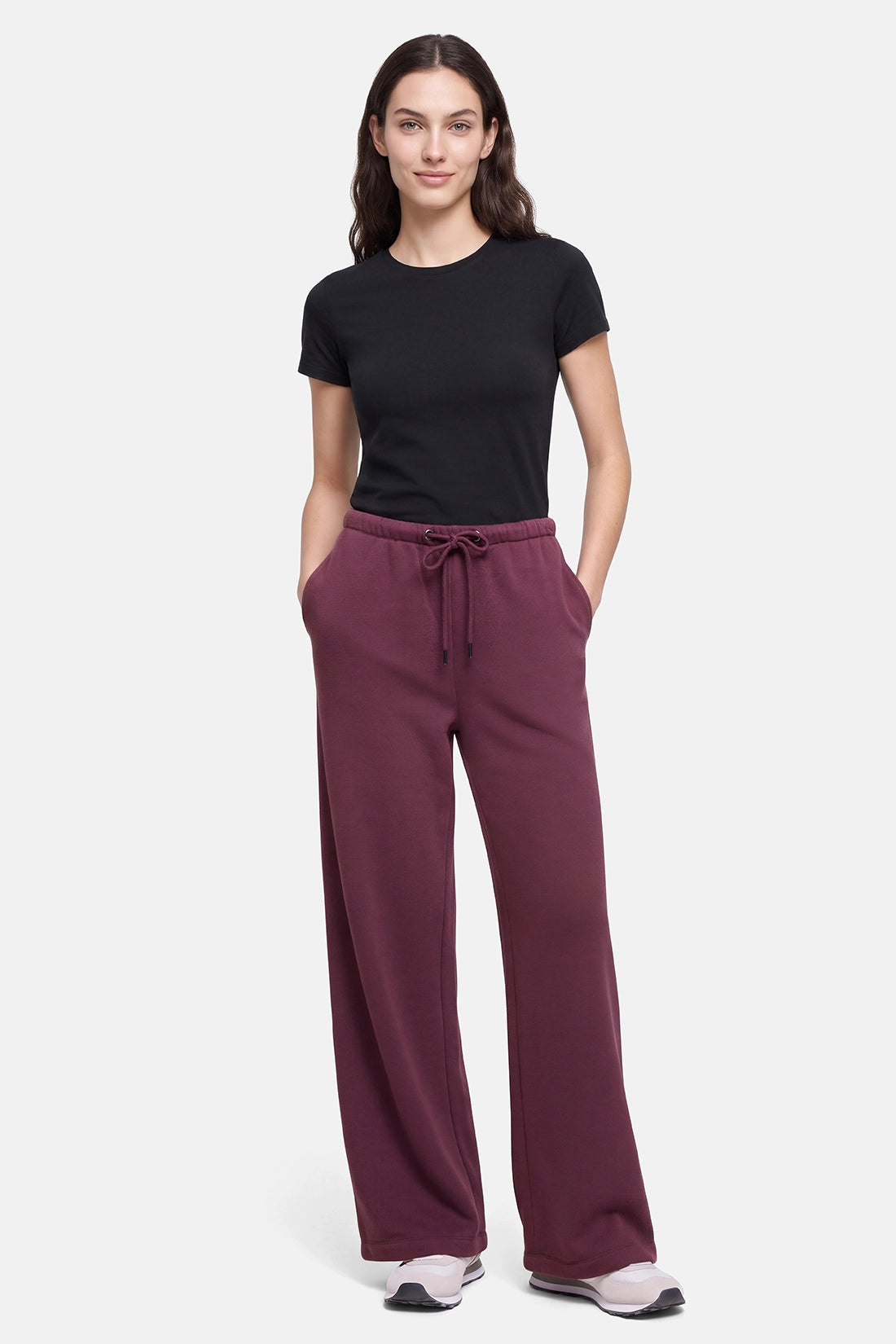A woman modeling a pair of wide-leg sweatpants in a deep burgundy color, paired with a black t-shirt and white sneakers, against a light gray studio background. 