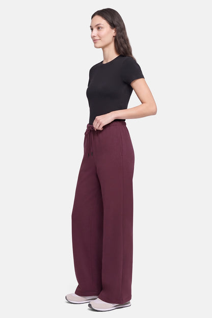 A woman modeling a pair of wide-leg trousers in a deep burgundy color, paired with a black short-sleeve top and light gray sneakers, shown from a side profile against a light gray background. 