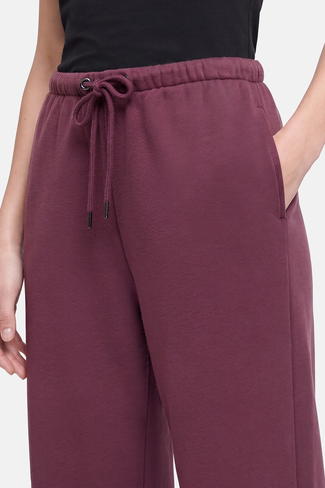 A close-up of a person wearing wide-leg sweatpants in a dusty plum or burgundy shade, paired with a black top. The sweatpants feature an elastic waistband with a matching drawstring and side pockets. 