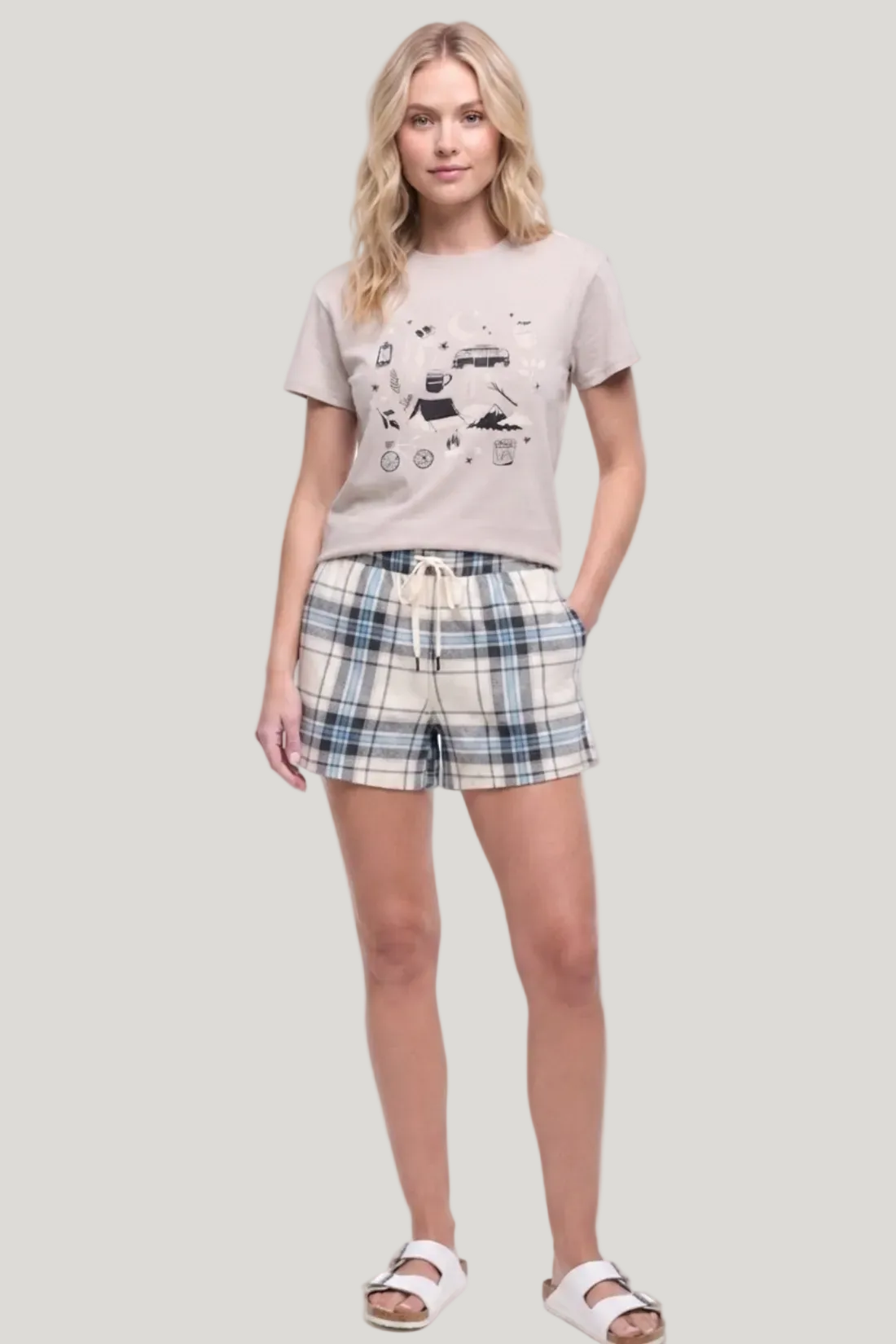 A woman modeling a two-piece lounge set consisting of a light beige graphic t-shirt and matching plaid shorts in beige, blue, and white, paired with white slide sandals on a white studio background.