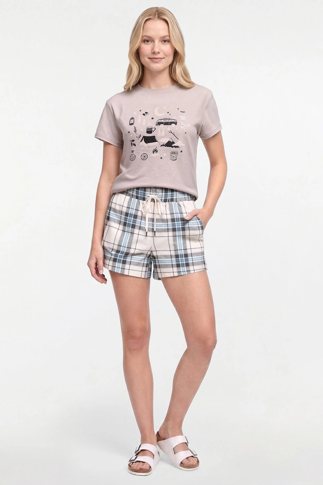 A woman modeling a two-piece lounge set consisting of a light beige graphic t-shirt and matching plaid shorts in beige, blue, and white, paired with white slide sandals on a white studio background.
