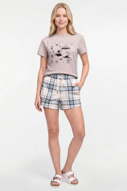 A woman modeling a two-piece lounge set consisting of a light beige graphic t-shirt and matching plaid shorts in beige, blue, and white, paired with white slide sandals on a white studio background. 
