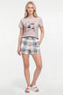 A woman modeling a two-piece lounge set consisting of a light beige graphic t-shirt and matching plaid shorts in beige, blue, and white, paired with white slide sandals on a white studio background. 