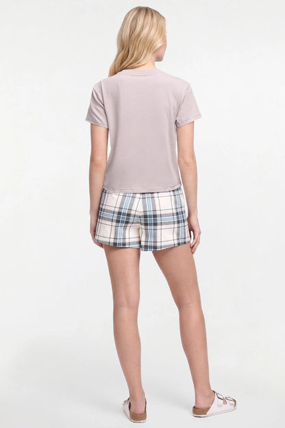 A woman shown from the back wearing a light taupe short-sleeve t-shirt and plaid shorts in white, blue, and gray, paired with white slide sandals, against a plain white studio background.