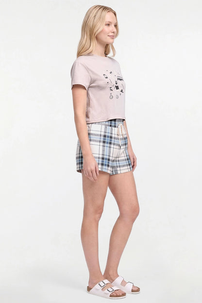 A woman modeling a light grey-tan cropped t-shirt with a black graphic print, paired with blue and white plaid shorts and pink double-strap sandals, against a plain white studio background.