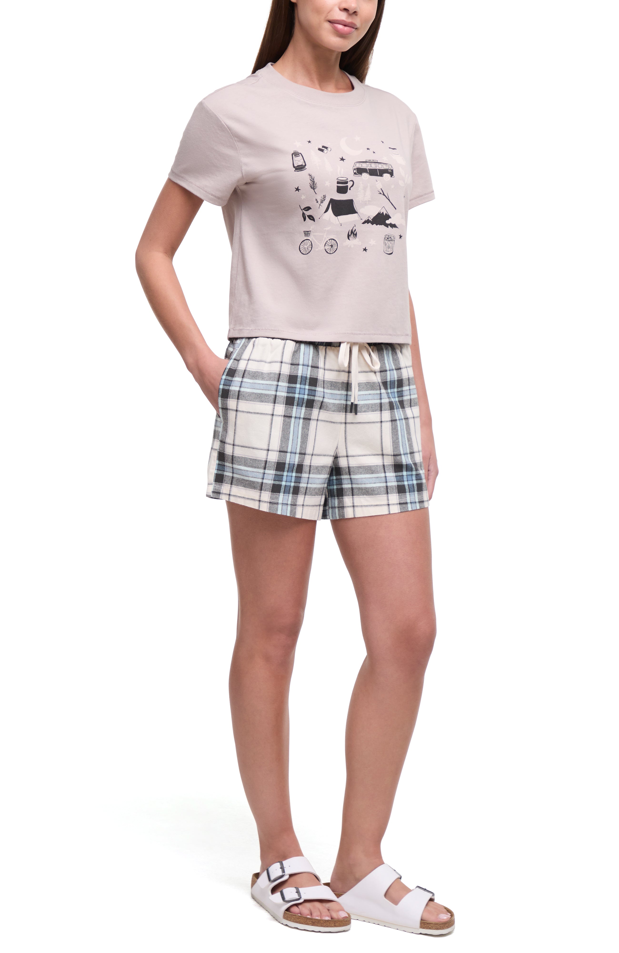 A woman modeling a two-piece lounge set consisting of a light beige graphic t-shirt and matching plaid shorts in beige, blue, and white, paired with white slide sandals on a white studio background.