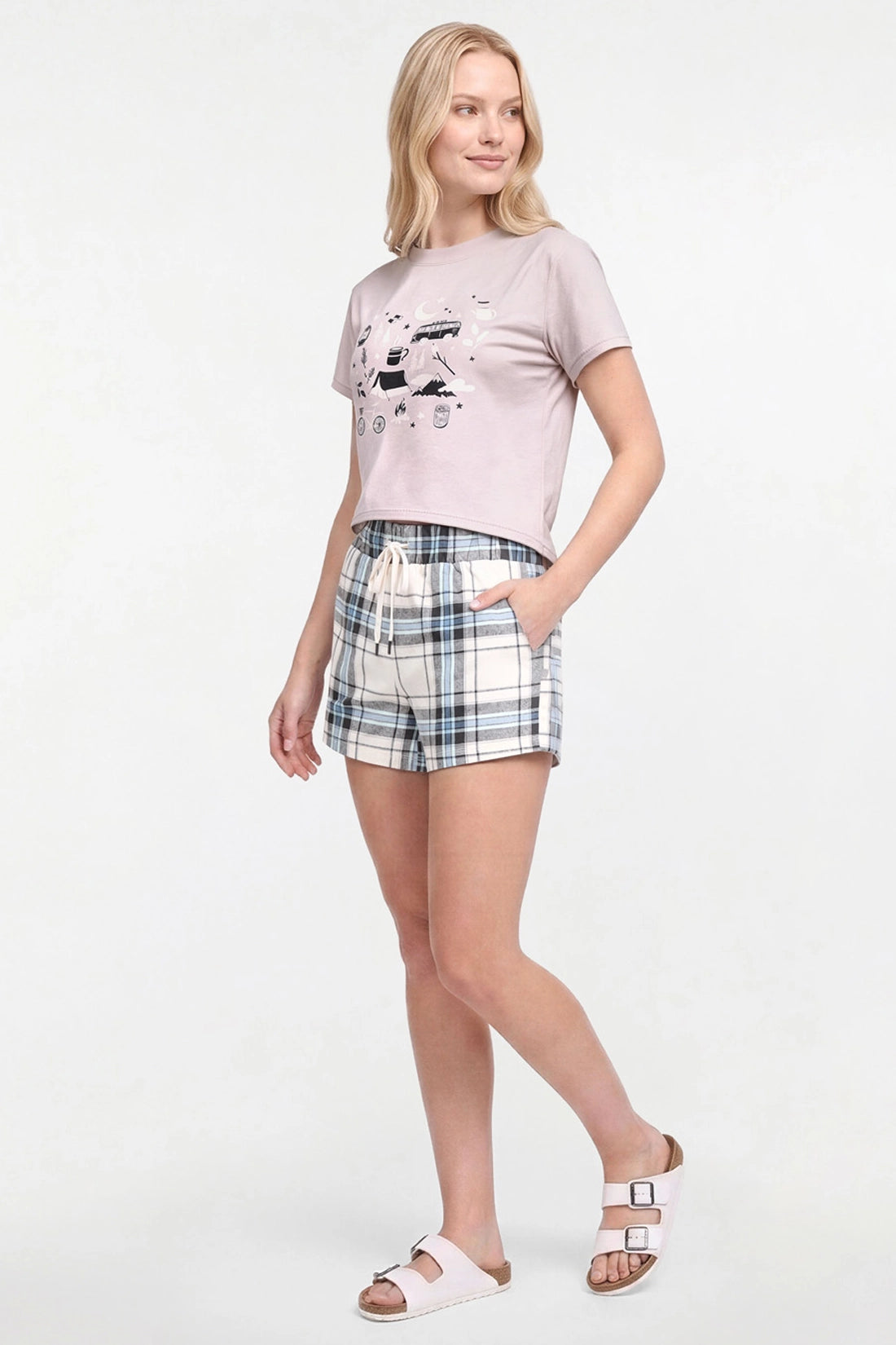 A woman modeling a two-piece lounge set consisting of a light beige graphic t-shirt and matching plaid shorts in beige, blue, and white, paired with white slide sandals on a white studio background.