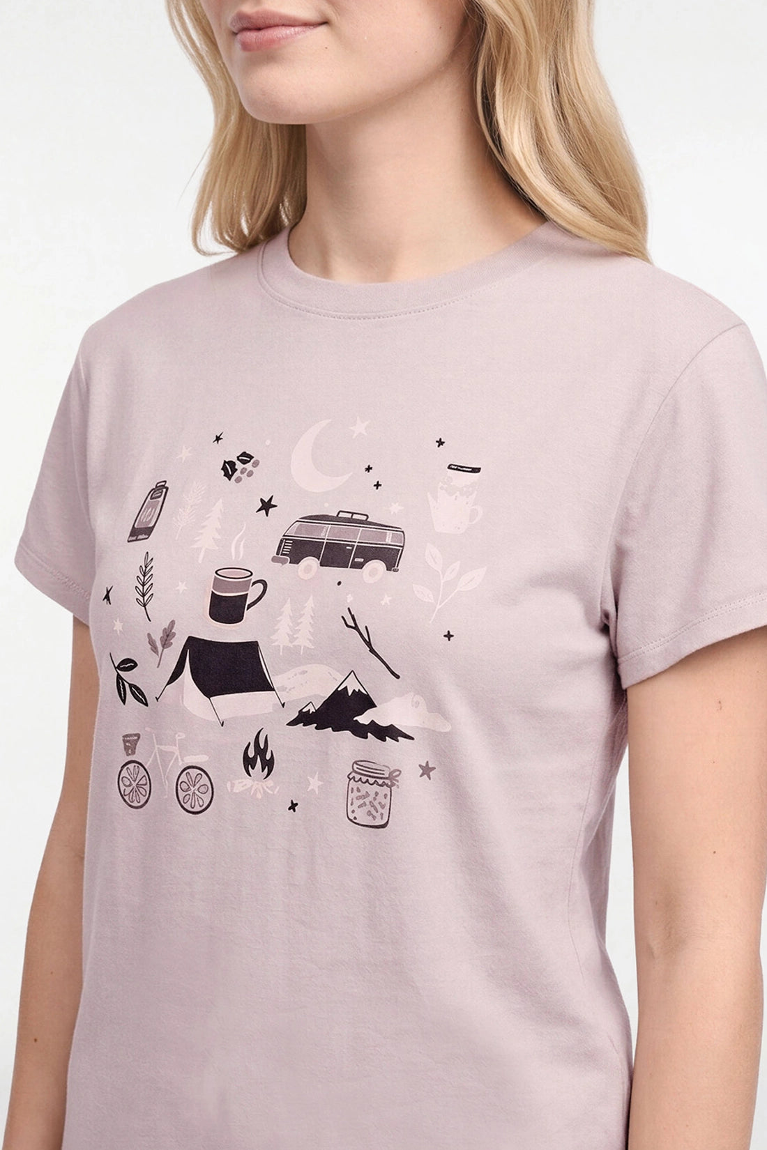 A woman wearing a light pink crewneck t-shirt with a black camping-themed graphic print on the front, featuring a van, tent, campfire, and stars.