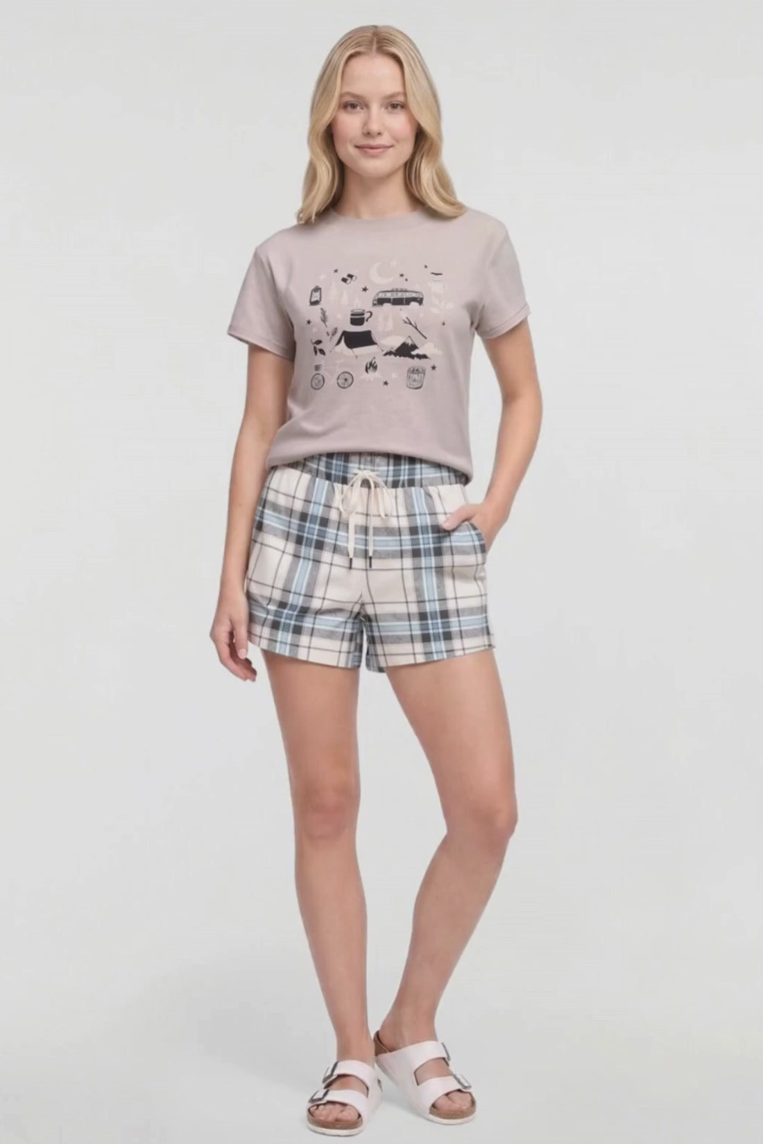 Video of a woman modeling a two-piece lounge set consisting of a light beige graphic t-shirt and matching plaid shorts in beige, blue, and white, paired with white slide sandals on a white studio background. #color_atmosphere