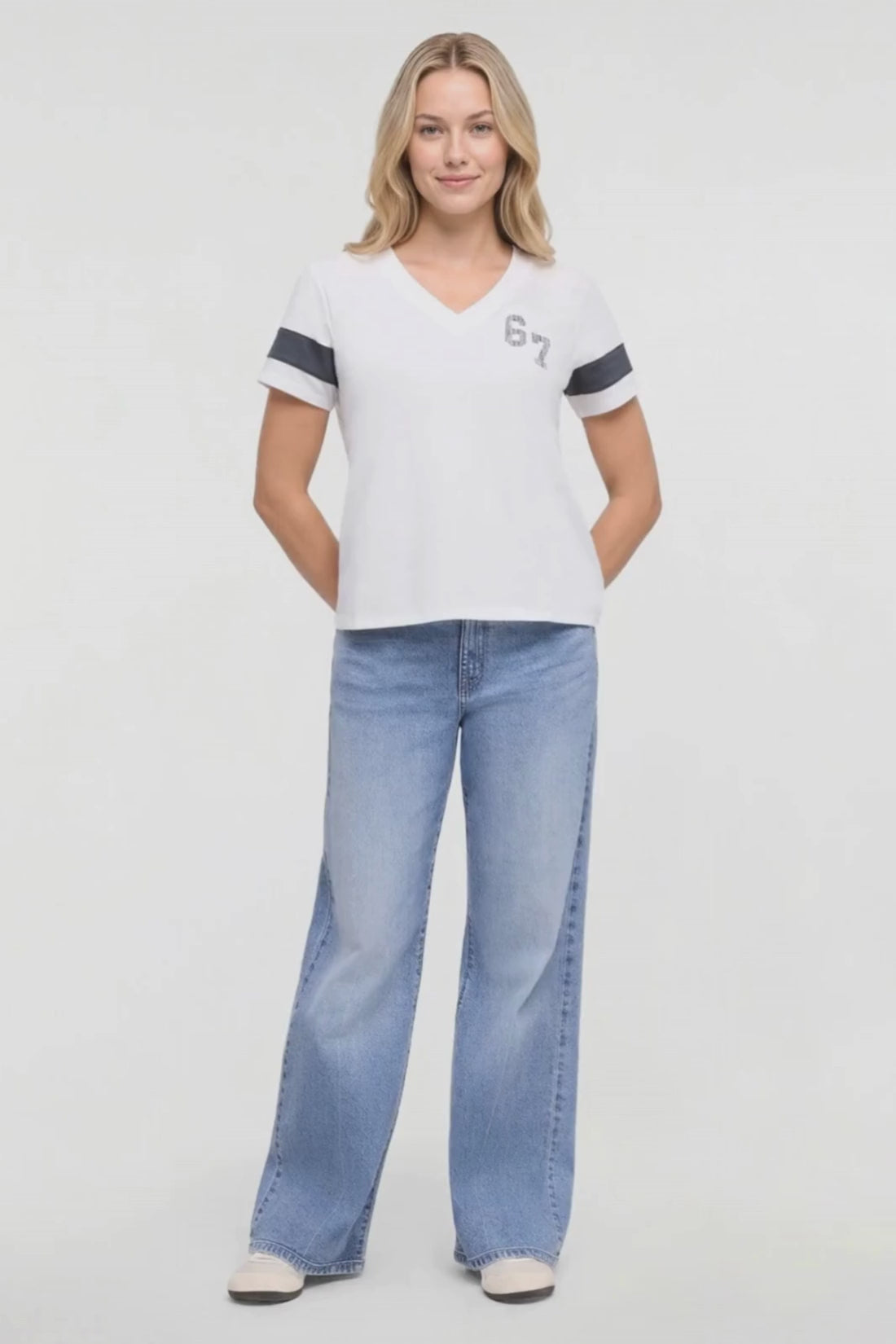 Video of a woman wearing a white v-neck t-shirt with dark gray sleeve stripes and a gray '67' graphic on the chest, paired with blue jeans, against a plain white background. #color_snow-white