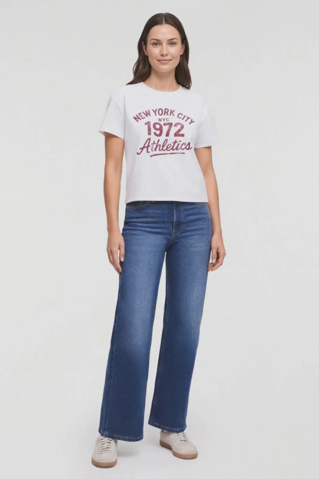 Video of a woman modeling a white short-sleeve graphic T-shirt with red text reading 'New York City NYC 1972 Athletics' and blue jeans, standing against a plain light background. #color_snow-white