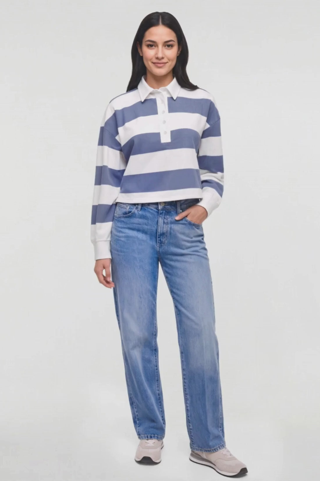 Video of a woman modeling a blue and white striped long-sleeve rugby shirt with light-wash blue jeans and light gray sneakers, standing against a plain white studio background. 