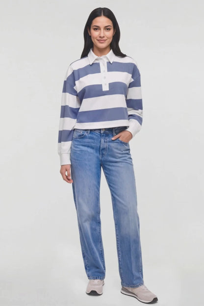Video of a woman modeling a blue and white striped long-sleeve rugby shirt with light-wash blue jeans and light gray sneakers, standing against a plain white studio background. 