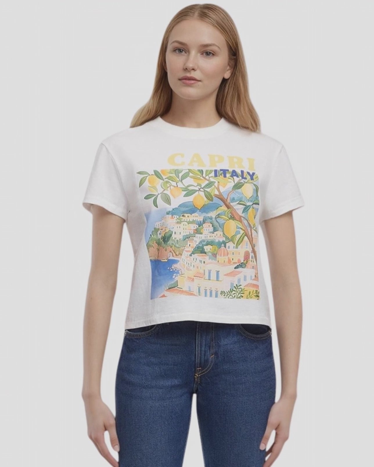 Video view of woman wearing a white t-shirt with a colorful graphic design of Capri, Italy, featuring buildings, trees, and lemons and 'CAPRI ITALY' text and blue jeans on a plain background. #color_snow-white
