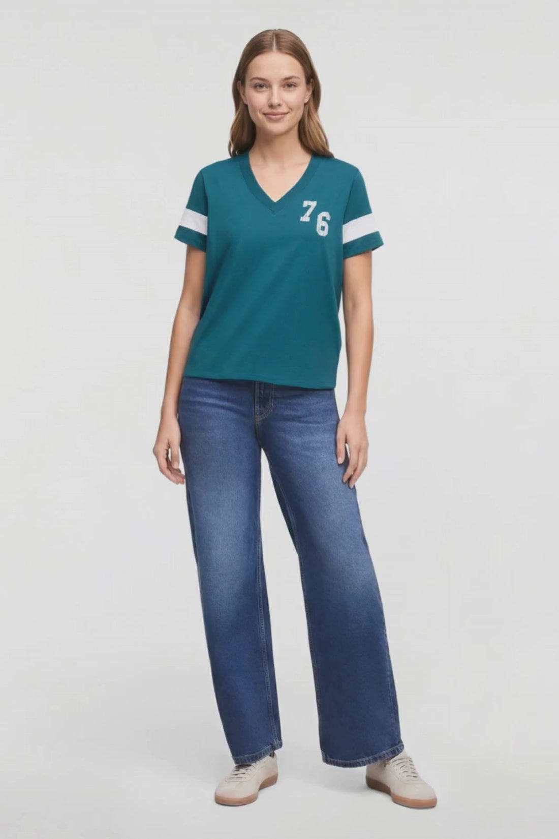 Video of woman modeling a teal V-neck short-sleeve t-shirt with white stripes on the sleeves and a white logo on the chest, paired with blue jeans and beige sneakers, against a plain white studio background. 