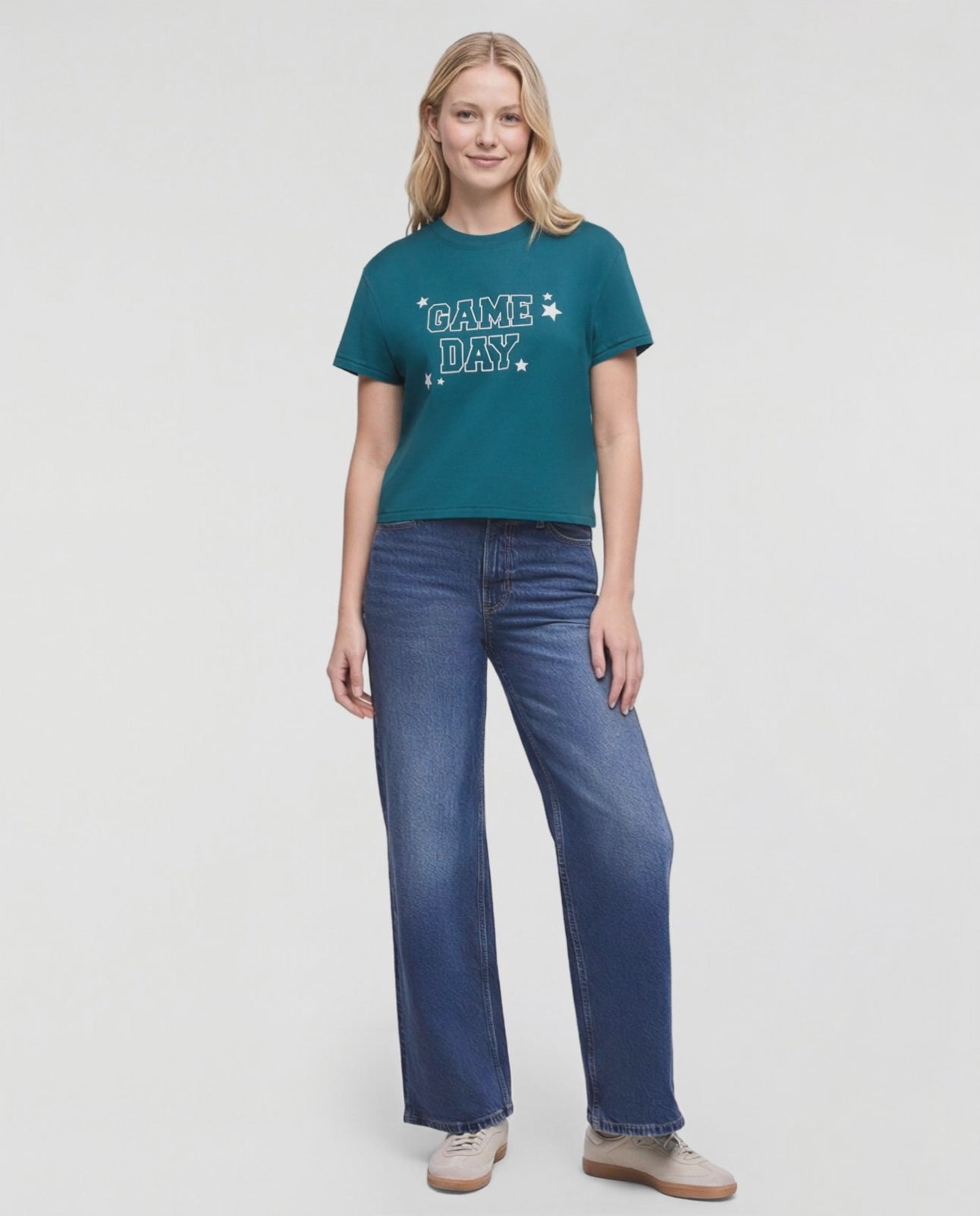 Video of a woman modeling a teal short-sleeve graphic t-shirt with 'GAME DAY!' printed in white on the chest, paired with wide-leg blue jeans and beige sneakers, against a plain white studio background. #color_deep-teal
